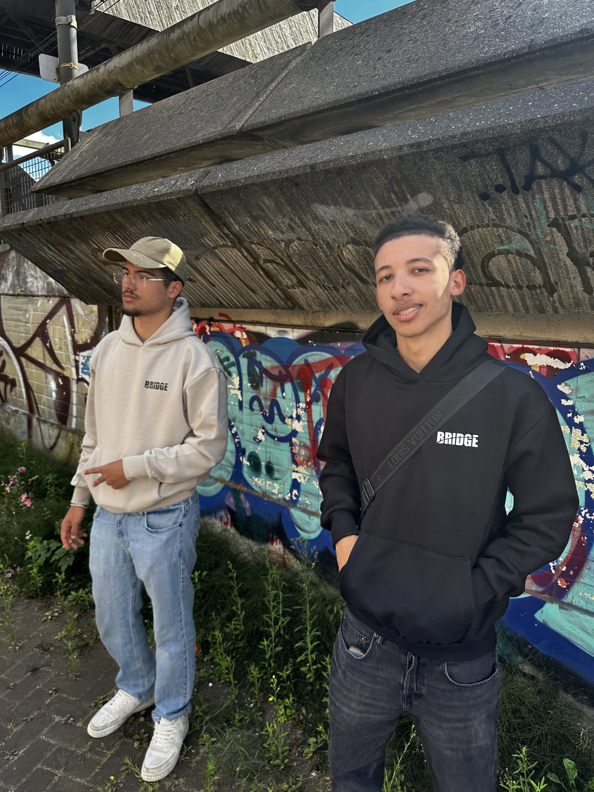 Twee modellen dragen BRIDGE hoodies in zwart en beige, verkrijgbaar in maat M, L en XL