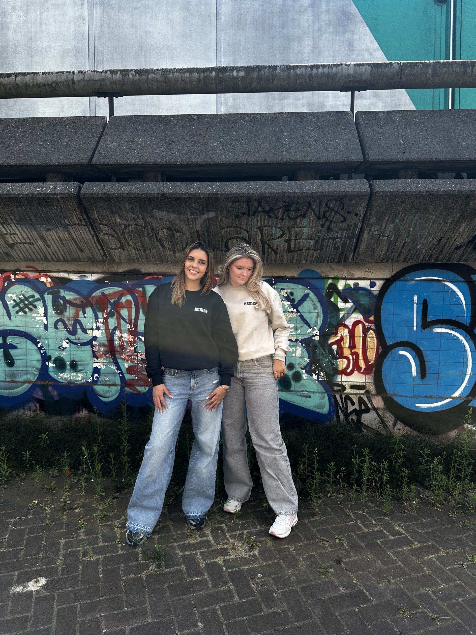 Duo foto met BRIDGE sweaters in zwart en beige, exclusief design van kunstenaar Luuk Bode, unisex streetwear in maat M, L en XL