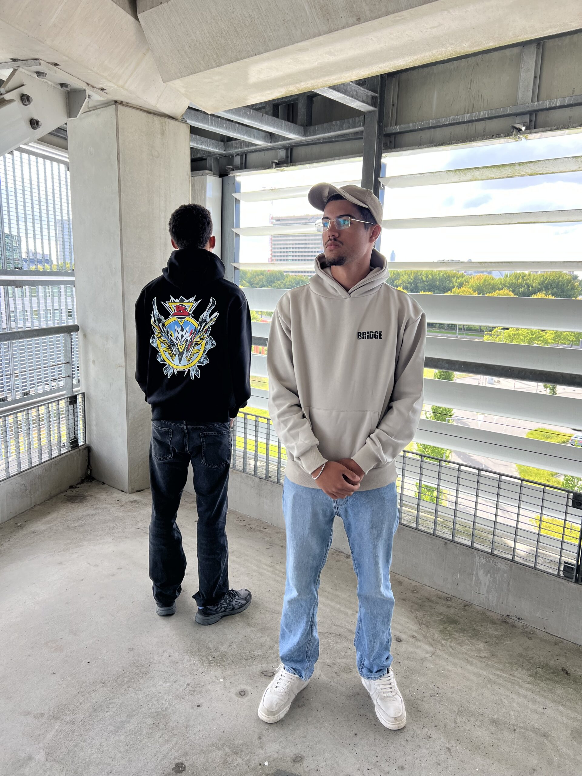 Twee modellen dragen BRIDGE hoodies in zwart en beige, exclusief design ontworpen door Luuk Bode, verkrijgbaar in maat M, L en XL